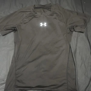 Svart compression-shirt från Under Armour - Svart kortärmad compression-shirt från Under Armour med logga på bröstet. Tillverkad i ett lätt och stretchigt syntetmaterial som andas, perfekt för träning. Snygg och sportig passform som sitter skönt på kroppen.