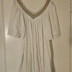 Vit strandklänning  - Luftig vit strandklänning med v-ringning och breda ärmar. Klänningen har ett guldfärgat broderi längs halsringningen. Onesize men lösare modell som nog passar många 