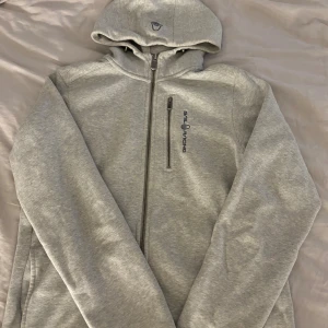 Grå hoodie från Sail Racing - Snygg ljusgrå hoodie från Sail Racing med hel dragkedja och bröstficka med dragkedja. Tröjan har broderad logga på bröstet och en skön huva. Som ny och inga märken eller fläckar. Passar en som har xs/s