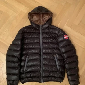 Svart Colmar pufferjacka med huva - Snygg svart pufferjacka från Colmar. Jackan har två dragkedjefickor framtill, quiltad design och tydliga Colmar-loggor på bröstet och ärmen. Perfekt för kalla dagar och riktigt skön att bära. Ett litet hål i innerfickan men resten av skicket är mycket bra utan att det finns några små hål som är igen sydda! Storleken i jackan är 50=M