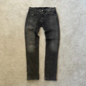 Replay Anbass grå/svarta jeans, W29 L32 - Snygga Replay Anbass jeans i svart tvätt med diskret slitning. Modellen har smal passform, klassiska fem fickor och dragkedjegylf. Jeansen är tillverkade i stretchig denim för extra komfort och har Replay-logga på fickan.