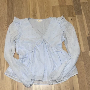 Ljusblå volangblus från Gina Tricot - Supersöt ljusblå blus från Gina Tricot med volangdetaljer på axlar och omlott framtill. Blusen har diskret prickigt strukturmönster, v-ringning och långa ärmar med lätt puff. Perfekt för dig som gillar romantisk och trendig stil. Passar mos om vanligtvis är en storlek xs/s