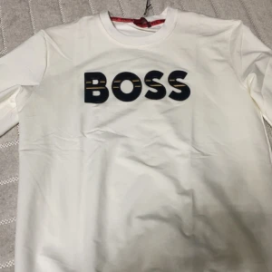 Vit långärmad t-shirt från Hugo Boss - Vit långärmad t-shirt från Hugo Boss med stor svart BOSS-logga framtill. Klassisk rund halsringning och stilren design. Tillverkad i mjuk bomull för en skön känsla. Perfekt för dig som gillar clean och exklusiv streetstyle. Storlek L men passar M