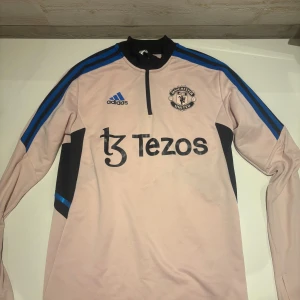 Manchester United träningströja XS - Ljusrosa långärmad träningströja från Adidas x Manchester United med svarta och blå detaljer, halv dragkedja och klubbmärke på bröstet. Perfekt för fotbollsträning eller chill dagar. Materialet är polyester och modellen har en sportig passform.