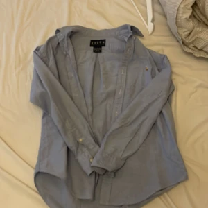 Ljusblå skjorta från Ralph Lauren - Klassisk ljusblå skjorta från Ralph Lauren i 100% bomull. Skjortan har lång ärm, knappar framtill och den ikoniska broderade polospelaren på bröstet. Perfekt oversized fit för en chill och stilren look.