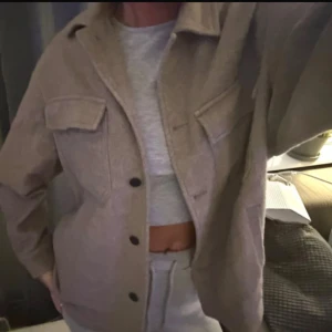 Beige overshirt från Zara i ullmix - Overshirt från Zara i beige ton med stora svarta knappar och två bröstfickor med lock. Jackan har en avslappnad passform och är gjord i en mjuk ullmix som passar perfekt till lager-på-lager. Snygg att styla med både jeans och mjukisbyxor. Endast använd några fåtal gånger, har inga defekter! 