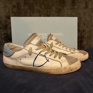 Philippe Model Junior  - Snygga vita Philippe Model Junior sneakers i skinn med beige detaljer och blå logga på sidan. Skorna har snörning, platt sula och en ljusblå häl med röd söm. Perfekta för dig som gillar stilrena men ändå unika sneakers.