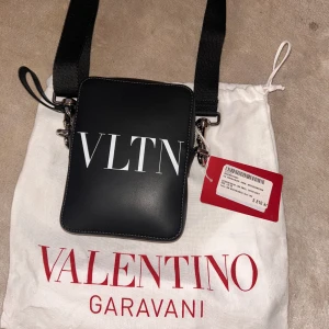 Svart axelväska Valentino Garavani (Köpt För 5800Dkk) - Snygg svart axelväska från Valentino Garavani med bred axelrem och silverfärgade metalldetaljer. Väskan har VLTN-tryck på framsidan och logga på baksidan. Tillverkad i slätt skinn och levereras med dustbag och tags. 