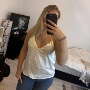 Vit satinlinne med smala axelband - Säljer ett stilrent vitt linne i satin med smala axelband som korsas i ryggen. Toppen har v-ringning framtill och en lätt glansig finish som ger en lyxig känsla. Perfekt att matcha med jeans eller kjol för en snygg look.