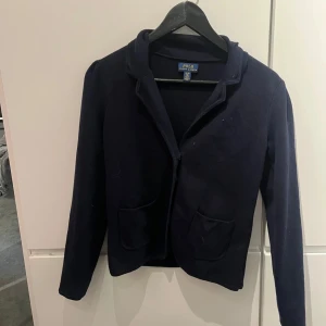 Mörkblå kavaj från Polo Ralph Lauren - Snygg mörkblå kavaj från Polo Ralph Lauren med knappar framtill och två fickor. Kavajen har långa ärmar och en klassisk krage. Kavajen har inte används så mycket, bara några gånger, och den är i bra skick 