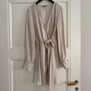 Beige omlottklänning med knyt - Superfin beige kort omlottklänning med lång ärm och knyt i midjan. Klänningen har v-ringning och är gjord i ett mjukt, lätt bomullstyg med diskret struktur. Perfekt för dig som gillar en enkel men stilren look.