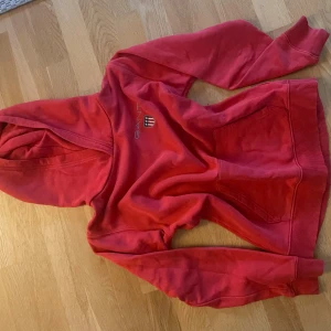 Röd hoodie från GANT - Säljer en röd hoodie från GANT med broderad logga och emblem på bröstet. Tröjan har huva, magficka och ribbade muddar vid ärmslut och nederkant. Perfekt för dig som gillar klassisk och sportig stil.