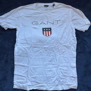 Vit t-shirt från GANT med logga - Vit t-shirt från GANT. Klassisk rund halsringning och korta ärmar. Perfekt för en clean och stilren look. Fått den i present men är aldrig använd. Storleken är 176 cm (16 år) men sitter som en S/M. Fler bilder kan skickas om önskas och pris går att diskutera.