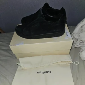 Svarta sneakers från Axel Arigato - Stilrena svarta sneakers från Axel Arigato i mocka med diskret logga på sidan. Skorna har en klassisk låg profil och platt sula, perfekta för en clean och modern look. Kommer med originalpåse och låda.