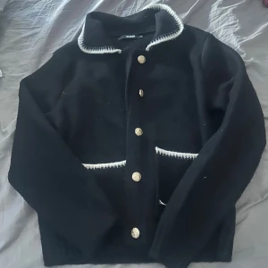 Svart cardigan jacka från Bik Bok - Svart cardigan jacka från Bik Bok med guldknappar och vita detaljer vid krage och fickor. Jackan har lång ärm, klassisk krage och två fickor framtill. Perfekt för dig som gillar en stilren och chic look.