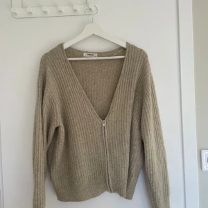 Beige ribbad kofta från MSCH - Säljer en beige ribbad kofta från MSCH med v-ringning och dragkedja framtill. Koftan är stickad och har långa ärmar, perfekt för lager på lager under höst och vinter. Enkel och stilren design som passar till det mesta.