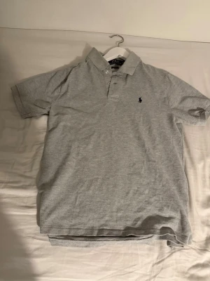Grå pikétröja från Polo Ralph Lauren - Säljer en klassisk grå pikétröja från Polo Ralph Lauren i storlek L/G. Tröjan har korta ärmar, krage och knappar framtill. Den är i bomull och har den ikoniska lilla broderade loggan på bröstet. Perfekt för en chill och stilren look.
