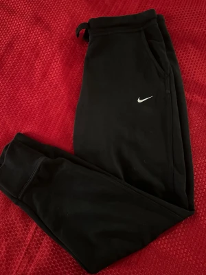 Dam Svarta mjukisbyxor från Nike storlek S - Svarta mjukisbyxor från Nike med klassisk logga på benet. Byxorna har snörning i midjan och ribbade muddar vid bensluten. Tillverkade i mjukt bomullsmaterial som är skönt mot huden. Perfekta för chill eller träning.