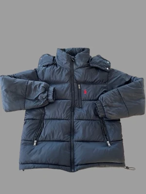 Svart pufferjacka från Polo Ralph Lauren - Säljer en svart pufferjacka från Polo Ralph Lauren med hög krage och dragkedja framtill. Jackan har två fickor med dragkedja, elastiska muddar och den klassiska röda loggan på bröstet. Perfekt för kalla dagar och riktigt snygg till alla outfits.