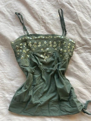 Grön paljettprydd linnetopp - Snygg grön linnetopp med tunna axelband och dekorativa paljetter i guld på  bröstpartiet. Toppen har spetsdetaljer och lätt rynkade sömmar framtill för en feminin look. Perfekt för dig som vill sticka ut lite extra. Går att knyta åt i höfterna🩷