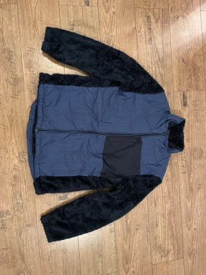 Blå teddyjacka från Nautica, L - Säljer en blå teddyjacka från Nautica i storlek L. Jackan har svarta fluffiga teddyärmar och insida, hög krage och dragkedja framtill. Utsidan är blå med en svart bröstficka med dragkedja. Jackan är knappt använd och har inga skador på sig.