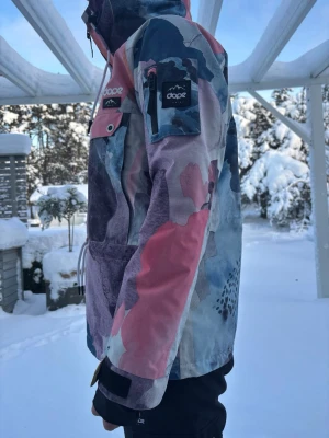 Flerfärgad vindjacka från Dopesnow - Säljer en riktigt snygg skidjacka från Dope med abstrakt mönster i blått, rosa, lila och vitt. Jackan har en stormsäker huva som funkar lika bra med hjälm som utan. Den har ventalation under ärmarna som passar perfekt vid en varmare skiddag. På vänster arm finns ens ficka perfekt för liftkort. Jackan har en stor magficka med dragkedja och snörning i midjan. Den har handelsmuddar som är stretchiga och bekväma.  Perfekt för dig som vill sticka ut i backen och samtidigt hålla dig varm.  