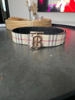 Rutig Burberry bälte med BB-spänne - Säljer ett klassiskt Burberry bälte med ikoniskt rutmönster i beige, svart, vitt och rött. Bältet har även en hel svart sida och är 110 cm