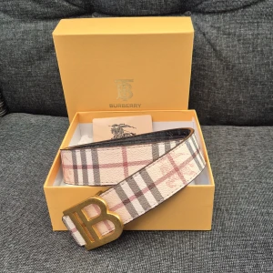 Rutig Burberry bälte med guldspänne 105 cm - Säljer ett klassiskt Burberry bälte i beige och brunt med det ikoniska rutmönstret och ett stort B-format guldspänne. Bältet är tillverkat i skinn och levereras i originalask med dustbag och certifikat. Perfekt accessoar för att lyfta din outfit.