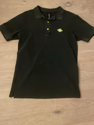 Svart Replay henley med neonknappar - Svart henley-skjorta från Replay med korta ärmar och krage. Snygga neonfärgade knappar och Replay-logga i gult på bröstet. Materialet är mjuk bomull och passformen är normal. Perfekt för dig som gillar stilrena detaljer och vill sticka ut lite extra.