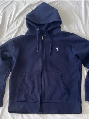 Ralph lauren zip hoodie - Tja säljer en ralph lauren zip hoodie då den börjar bli för lite för mig. Det är en klassisk marinblå hoodie med hel dragkedja och en vit Polo-logga på bröstet. Tidlös modell som funkar lika bra till vardags som för en mer avslappnad stil. Mjuk och bekväm med huva, justerbara snören och två framfickor. Den har en liten fläck på baksidan av ärmen (se 3 bild)