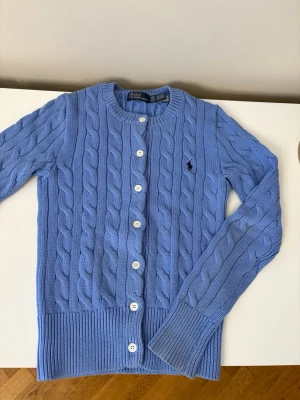 Blå kabelstickad kofta  - Supersnygg blå kabelstickad kofta från Polo Ralph Lauren. Klassisk modell med rund hals, vita knappar framtill och broderad logga på bröstet. 
