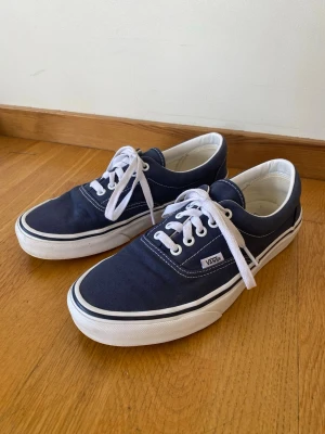 Marinblåa Vans sneakers i storlek 40 - Säljer ett par klassiska marinblåa Vans sneakers med vita snören och vit sula. Skorna har rund tå, lågt skaft och är tillverkade i bomullstyg med kontrastsömmar. Skorna är i bra skick och säljes eftersom de är för små nu. Hör gärna av dig vid eventuella frågor.