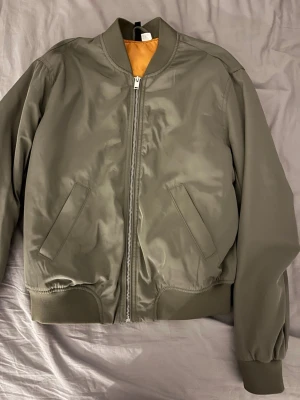 Olivgrön bomberjacka med orange foder - Snygg olivgrön bomberjacka med klassisk ribbad krage, muddar och midja. Jackan har orange foder, två snedställda fickor framtill och dragkedja i silver. Tillverkad i ett glansigt syntetmaterial som ger en modern look. Perfekt för dig som vill ha en stilren och enkel jacka.
