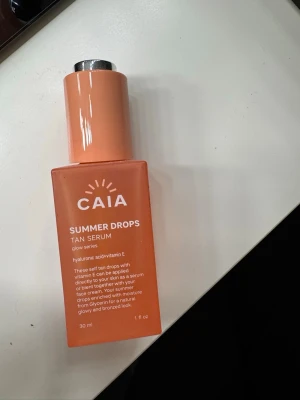 CAIA Summer Drops Tan Serum - Glow-serum i en snygg orange glasflaska med silvrig kork. Innehåller hyaluronsyra och vitamin E för återfuktning och lyster. Perfekt för att blanda med ansiktskräm eller använda direkt på huden för en solkysst look.
