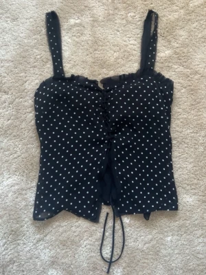 Svart prickig korsettopp från Zara - Supersöt svart korsettopp från Zara Studio med vita prickar och snörning framtill. Toppen har tunna axelband, volangkant upptill och är figurnära i modellen. Perfekt för dig som vill ha en trendig och lekfull look.