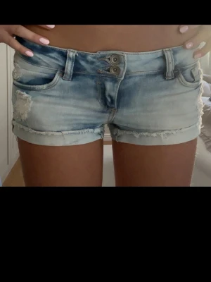 Lågmidjade ljusblå jeansshorts från Tally Weijl - Säljer ett par ljusblå jeansshorts från Tally Weijl med slitna detaljer och fransig kant. Shortsen har låg midja, dubbla knappar framtill och klassiska fem fickor. Perfekta till sommaren och riktigt snygga till sneakers eller sandaler.