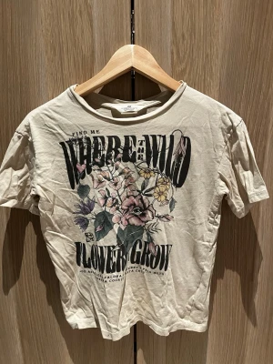 Beige blommig t-shirt från H&M - Beige t-shirt från H&M med trycket 'WHERE THE WILD FLOWERS GROW' och ett stort blommotiv i pastellfärger på framsidan. sopig passform och klassisk rund hals. Perfekt för dig som gillar vintagevibbar och coola prints.