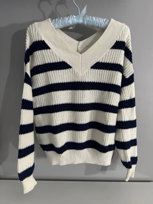 Randig stickad tröja med v-ringning - Säljer en ribbstickad tröja med bred v-ringning och marinblå och vita ränder. Tröjan har lång ärm och en oversized känsla, perfekt för lager på lager. Klassisk och enkel design som funkar till mycket.