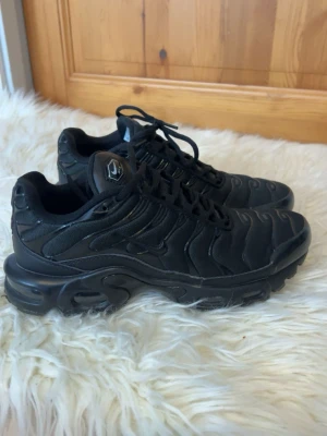 Nike Air Max Plus helsvarta sneakers - Säljer ett par Nike Air Max Plus i helsvart färg. Skorna har en chunky sula med synlig Air-enhet, mesh och syntetpaneler samt klassisk Tn-detalj på hälen. Snygg vågig design och snörning framtill. Perfekta för dig som gillar streetwear och sportig stil.