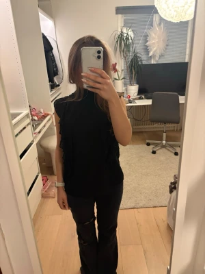 Svart blus med volanger och prickar - Säljer en svart blus med hög krage och volangdetaljer längs axlarna. Blusen är ärmlös och har ett diskret prickigt mönster.