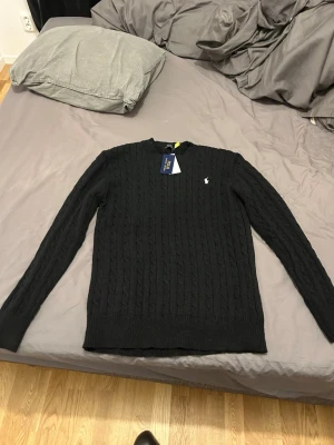 Svart kabelstickad tröja Polo Ralph Lauren - Klassisk svart kabelstickad tröja från Polo Ralph Lauren med rund halsringning och diskret broderad logga på bröstet. Långärmad modell i mjuk ullblandning som passar perfekt till jeans eller chinos. Aldrig använd då den ej passat mig