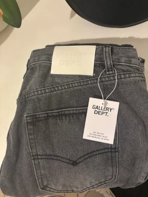 Grå jeans från Gallery Dept. - Säljer ett par grå jeans från Gallery Dept. med råa, fransiga benslut och klassisk femficksdesign. Jeansen har en rak passform och är tillverkade i slitstarkt bomullsmaterial. Stora vita patchar med logga bak i midjan och på insidan.