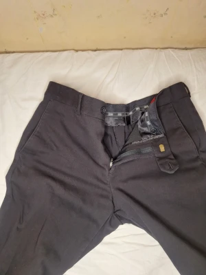 Svarta kostymbyxor från Corleani - Snygga svarta kostymbyxor från Corleani med klassisk rak passform. Byxorna har bälteshällor, framfickor och bakfickor med knapp. Materialet är en mix av bomull, nylon och lycra för skön stretch och stilren look. Perfekta till skjorta eller kavaj.