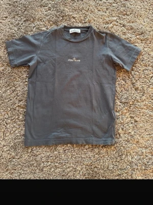 Mörkgrå Stone Island Junior t-shirt 152 cm - Snygg mörkgrå t-shirt från Stone Island Junior med diskret logga på bröstet och coolt grafiskt tryck på ryggen. Klassisk passform och rund halsringning. Perfekt till streetwear-stilen.
