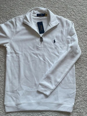 Vit half zip tröja från Polo Ralph Lauren - Säljer en stilren vit långärmad tröja från Polo Ralph Lauren med klassisk blå logga på bröstet. Tröjan har en snygg half zip-dragkedja och hög krage. Tillverkad i mjuk bomull för skön känsla och passar perfekt till lager på lager.
