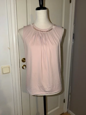 Ljusrosa blus med pärldetaljer - Säljer en ljusrosa blus i mjukt tyg med pärldetaljer runt halsringningen. Blusen är ärmlös och har fina veck framtill som ger ett elegant fall. Perfekt till både jeans och kjol för en snygg look.