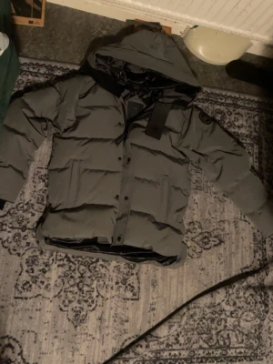 Canada goose macmillan Size L grå - Säljer en grå pufferjacka från Canada Goose med huva och svart logotyp på ärmen. Jackan har dragkedja och knappar framtill, samt en quiltad design som håller värmen. Perfekt för kalla dagar och levereras med original fodral. oanvänd 