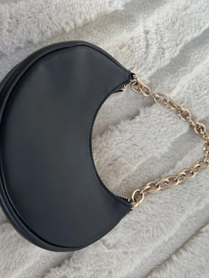 Svart axelväska med guldkedja - Säljer en svart axelväska i halvmåneform med en chunky guldfärgad kedja som axelband. Väskan är gjord i slätt syntetmaterial och har dragkedja upptill. Perfekt för dig som vill ha en stilren och trendig accessoar.