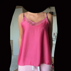 Rosa linnetopp med spetsdetaljer - Säljer en snygg rosa linnetopp med tunna axelband och spets längs v-ringningen. Toppen har en lös passform och är tillverkad i ett glansigt material som ger en lyxig känsla. Perfekt för dig som gillar en feminin och trendig stil.