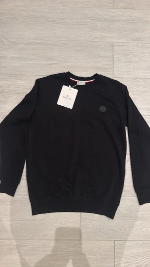 Svart sweatshirt från Moncler - Säljer en stilren svart sweatshirt från Moncler med rund hals och den perfekta logotyp på bröstet. Tröjan har långa ärmar och ribbade muddar vid ärmslut och nederkant. Materialet är mjuk bomull som ger en skön känsla. 